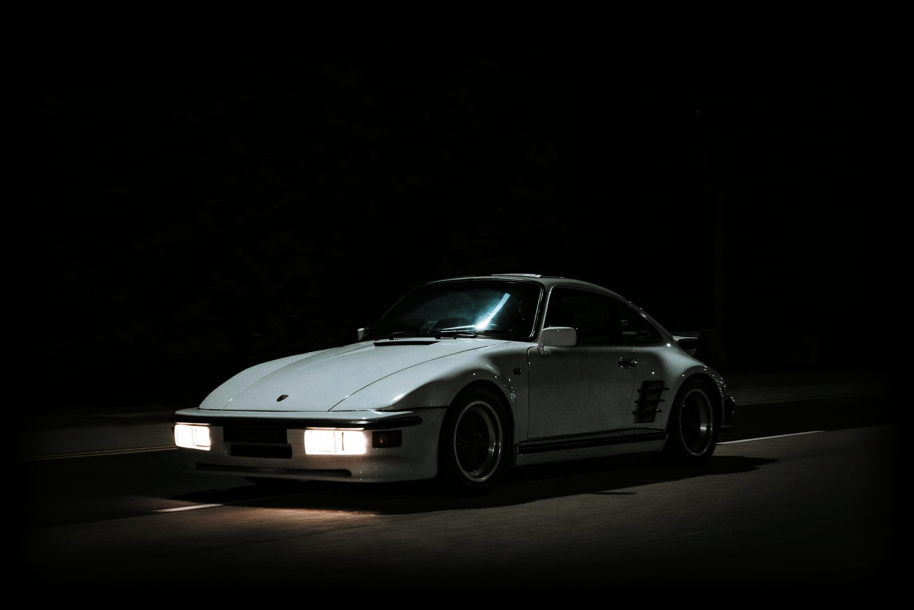 A white Porsche 911 with its headlights on, driving through the dark